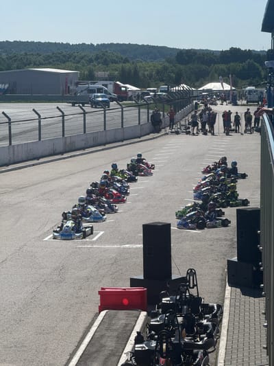 In Sekiotovo, Russia, a go-kart racing event is underway on a sunny day. The scene captures a group of young racers, clad in colorful helmets and racing suits, lined up on the asphalt track in their go-karts. The track features white lane markings, indicating a start or grid formation. In the background, a wooded landscape stretches across the horizon under a clear blue sky. Various tents and temporary structures suggest a race day atmosphere, with spectators and support personnel milling about. A fence separates the track from the spectator area, where more people are visible. Several vehicles, including vans and a truck, are parked in the vicinity. The overall impression is one of an organized sporting event taking place in an outdoor, semi-rural setting.