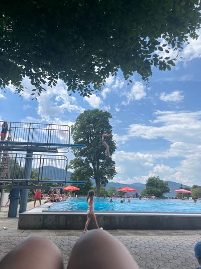 In Oberammergau, Germany, a diving board at an outdoor swimming pool is prominently featured. On the highest platform of the diving board, a person in a dark swimsuit is captured in mid-air, performing a dive. Below, the bright blue water of the pool is filled with people swimming and relaxing. The surrounding area has a paved walkway with people sitting, and a few red umbrellas provide shade. In the background, lush green trees and distant mountains under a partly cloudy sky complete the scenic view of this recreational facility.