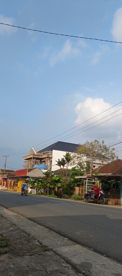 This is a vertical, eye-level, full-shot photograph taken on a sunny day in the city of Sukarame, Indonesia, depicting a residential street scene. The sky is a clear, pale blue with scattered, fluffy white clouds and traversed by thin, dark power lines. In the mid-ground, a road runs horizontally across the frame. Two people on a motorcycle, one driving and one passenger, are traveling away from the camera on the left side of the road. To the right of the road, a dark-colored scooter with a rider in a red garment is parked.
Several buildings are visible. On the far left, a row of single-story houses with tiled roofs and colorful facades are partially obscured by trees. Dominating the center and right of the mid-ground is a large, multi-story building under construction, its upper floors encased in scaffolding. It has a white exterior and a dark, tiled roof. Next to this, to the right, is a single-story house with a reddish-brown tiled roof and a green fence. Lush green tropical vegetation, including palm trees and flowering bushes, surrounds the houses and the construction site. The foreground shows the edge of the paved road and a concrete sidewalk with some cracks and patchy grass. The overall impression is of a developing residential area under a bright sky.