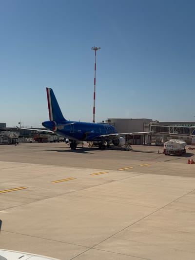 The image shows an ITA Airways aircraft parked at Falcone Borsellino Airport in Cinisi, Italy. The plane is positioned on the tarmac next to a gate, with a fuel truck and other airport vehicles nearby. A tall striped pole stands near the aircraft, and the airport terminal is visible in the background. The sky is clear and blue. The tarmac surface shows yellow painted lines. Part of another plane is visible in the bottom right of the frame. No people are clearly visible in the image.