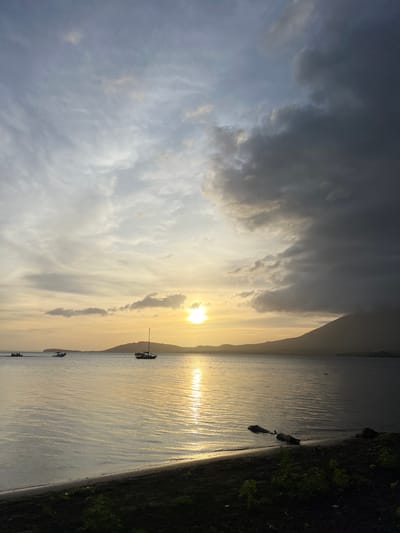 This is a photograph of a calm body of water, likely a bay or lake, with a distant shoreline and a mountain on the right. The sun is setting or rising, casting a warm golden light across the water and the sky. The sky is a mix of light and dark clouds, with the sun appearing as a bright, diffused orb.
In the water, there are three boats. Two smaller motorboats are closer to the left side of the frame, appearing to be traveling. Further to the right, a sailboat with a single mast is stationary or moving slowly. The shoreline in the foreground is dark and appears to be a beach or muddy bank, with some sparse vegetation. Two pieces of driftwood are partially submerged in the water near the shore. The overall scene is serene and evocative of a coastal or lakeside environment at dusk or dawn.