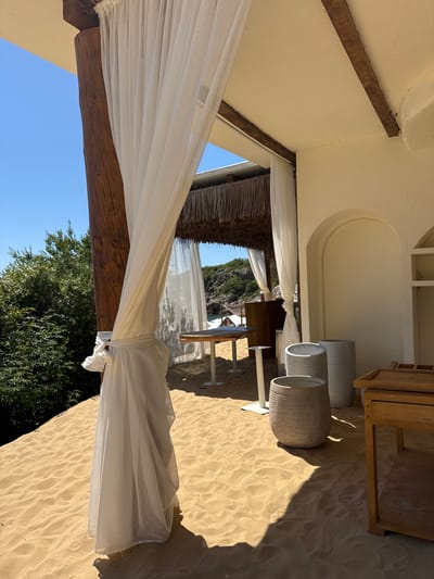 The image depicts an outdoor, partially covered area within an establishment in Vrba, Montenegro, characterized by a natural, rustic aesthetic. The ground is entirely covered in light beige sand, showing undulations and faint impressions. On the left, a large, dark-stained wooden pillar stands, wrapped with a sheer white fabric curtain tied in a knot. To the left of the pillar, dense green foliage, likely trees and bushes, thrives under a clear, bright blue sky.
The ceiling of the covered area is a textured, light beige, supported by visible dark wooden beams. In the mid-ground, another set of white sheer curtains hangs, partially obscuring a thatched roof structure further back. Beyond this, distant light-colored rocky terrain and a glimpse of blue water are visible.
To the right, a textured, light beige wall features two arched alcoves, one recessed and one appearing to be an opening or deeper niche. On the sand in front of this wall, three cylindrical planters or large vases of varying sizes and textures are positioned. One, closer to the foreground, exhibits a woven pattern. A light-colored wooden table with a white base is visible in the mid-ground, and another similar table is further back to the left. On the far right, a low, light-colored wooden table or cart with a slatted top and open shelving stands on the sand. Strong sunlight casts distinct shadows across the sand and objects. No people are present in the scene.