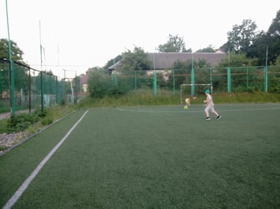 The image depicts an outdoor artificial turf soccer field in the city of Vyhoda, Ukraine. In the foreground, a white line runs diagonally across the field. In the middle ground, two individuals are present. One, wearing a light-colored t-shirt and shorts, along with a blue cap, is running to the right, facing away from the camera, with a soccer ball near his feet. To the left of this individual, another person, appearing smaller and wearing a yellow top, is also near a soccer ball. Behind the field, a green fence encloses the area, with trees and vegetation visible beyond it. In the background, a building with a dark, pitched roof, likely residential, is situated amidst more trees. The sky is overcast with a bright, pale hue.