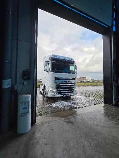 The image shows an exterior view from within a large, open industrial-style building located in Ostend, Belgium. A white DAF brand semi-truck tractor unit is positioned directly outside the open doorway, facing the camera. White foam and water cover the grid-patterned paver ground in front of and around the truck, indicating it is undergoing a washing process or has just been washed. The truck's headlights are illuminated, and its Belgian license plate, "1-YBB-702", is legible on the front bumper. The sky above is overcast with scattered clouds, and distant urban buildings alongside undeveloped land are visible in the background. Inside the building, to the left of the doorway, a light-colored Cligo water cooler/dispenser stands beside a grey pillar. A black electrical control box is mounted on the wall above the water cooler. The concrete floor inside the building is wet, with a blue light strip visible along the upper frame of the open doorway. No individuals are present in the scene.
