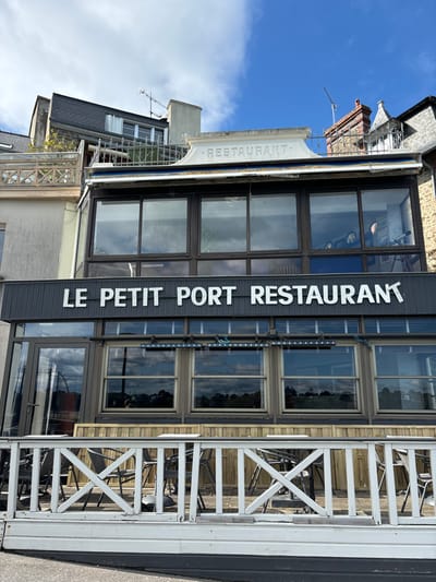 The image shows the "Le Petit Port Restaurant" located at 48.631, -2.052 in Dinard, France. The restaurant occupies a two-story building with a large sign above the entrance and numerous windows on the second floor. The first floor is mostly glass windows and doors that lead to a patio.  The patio is decorated with wooden tables and chairs.  The restaurant faces a white railing, partially obscuring the view of the street.  The sky is clear with some clouds, and the weather appears sunny.