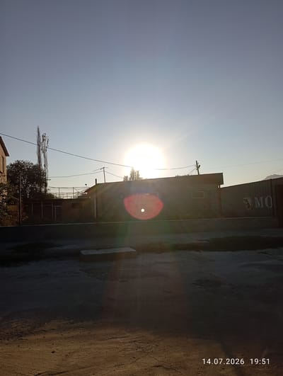 The image depicts a street scene at sunset in the city of Kyzyl-Sai, Kyrgyzstan. The sun is low on the horizon, creating a bright lens flare that obscures details in the sky and the surrounding buildings. The foreground is a dusty, unpaved street. In the middle ground, there are several low-rise buildings, some appearing to be made of brick and others of corrugated metal. One building has the partial inscription "MO" on its side. Trees and utility poles are also visible. The background consists of a clear sky and distant hills. The timestamp "14.07.2026 19:51" is visible in the bottom right corner. No people are present in the image.