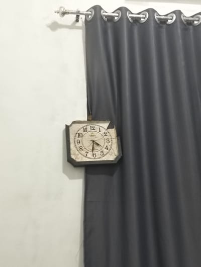 An analog clock with a square, white face and Roman numerals is hanging on a white wall to the left of a closed, dark gray curtain. The clock's face is aged and shows signs of wear. The hour hand points to the 4, and the minute hand points to the 7, indicating the time is approximately 4:35. Above the clock, a decorative metal curtain rod with finials is mounted on the wall. The curtain is made of a thick, textured material and hangs in vertical folds. The scene is indoors and appears to be a domestic setting.