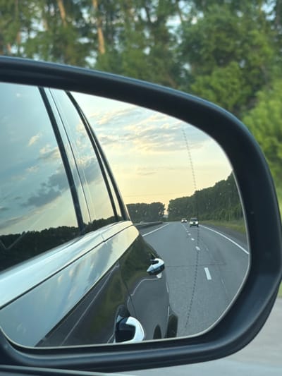 The image is a close-up, angled shot of a car's side-view mirror, reflecting a highway scene at dusk. The mirror is framed by the car's exterior, showing part of the vehicle's body and a window reflecting the sky and trees.
In the mirror's reflection, a two-lane highway stretches into the distance, bordered by dense green trees. The sky above is a pale yellow, with scattered, wispy clouds, indicative of sunset. Two cars are visible on the highway, moving away from the viewer. The car closest to the mirror is dark, with its headlights illuminated. The second car is further ahead and appears smaller. White lane markings are visible on the asphalt.
The environment appears to be rural or semi-rural, given the presence of the highway and the extensive tree line. The overall impression is one of travel during the evening. There is no indication of the location being within the city of Yemelyanovo, Russia. The scene is devoid of people.