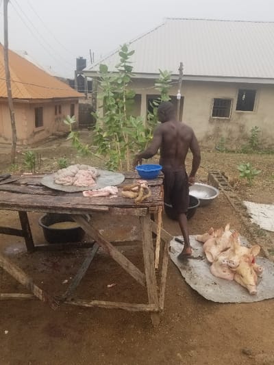 A man in a rural area of Pati Ridge, Nigeria, is butchering a pig. The man is standing next to a wooden table, on which sits a pig's carcass, along with other meat.  Several pig heads are laid out on the ground next to a metal sheet. There are two houses in the background, one with a brown roof and the other with a grey roof.  The ground is dirt and there are plants scattered around the scene.  The man is wearing shorts and no shirt. He is barefoot. The butchering appears to be taking place outside.  There is a blue plastic bowl on the table that contains the pig's internal organs.  There is also a black plastic bucket on the table which appears to be filled with water.  A metal bowl is sitting on the ground next to the pig's head.