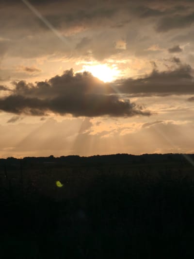 The image depicts a dramatic sunset over a rural landscape. The sky is filled with a mix of dark, ominous clouds and lighter, sun-drenched patches. Beams of sunlight pierce through the clouds, creating a crepuscular effect that illuminates the scene with an orange-gold hue. A silhouetted treeline or low hills forms the horizon. In the foreground, the landscape is almost entirely dark, appearing as a silhouette of vegetation, likely fields or tall grass. A faint, luminous yellow-green orb, possibly a lens flare or a reflection, is visible in the lower left portion of the foreground. The overall impression is one of natural beauty, with a hint of drama due to the contrasting light and shadow. The location in Barsukovskaya, Russia, suggests a vast, open expanse, typical of rural Russian countryside.