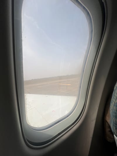 The view is from an airplane window. Outside the window is an airport runway. The sky is overcast. The location is unknown.