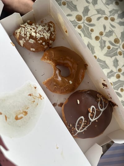 An overhead shot displays a white cardboard donut box containing three donuts and a white napkin with faint brown smudges. The donuts are arranged diagonally within the box. The top-left donut is half-eaten and covered in a caramel-colored glaze with white chocolate shavings. Adjacent to it, to the right, is a whole donut with the same caramel glaze, a small bite taken out of it. The bottom-right donut is fully intact, covered in dark chocolate glaze, decorated with a white icing swirl and chopped nuts. The background outside the box reveals a patterned surface, possibly a tablecloth, with a design of holly leaves and berries in grey and gold tones. The scene appears to be indoors, possibly in a home environment, with a person's arm visible at the top left of the frame. The location of Macclesfield, United Kingdom is not discernible from the image.
