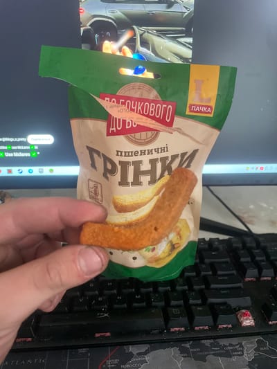 This is a close-up, slightly tilted shot of a hand holding a single, curved, golden-brown breadstick snack. The snack appears to be a russet-colored breadstick, possibly flavored with spices, with a slightly crispy texture. Behind the snack is a partially visible, open bag of "Грінки" (Grinki), which translates to croutons or rusks, made by the brand "ПАЧКА" (Pachka). The bag is predominantly green and beige with Cyrillic text and images of bread slices.
The background is a mixture of a computer monitor displaying what appears to be a web browser with a social media interface visible in the lower left, and a black mechanical keyboard. The monitor shows a social media feed with profile pictures and usernames, including "@things_x_pretty" and "use McLaren". The keyboard has red-accented keycaps, and part of a world map mousepad is visible on the right.
The lighting suggests an indoor setting, likely a room with artificial light, possibly a home office or living space. The overall impression is of someone taking a snack break while using their computer. There are no clear indicators of time of day or weather, and the emotional tone is neutral, focusing on the objects and text.