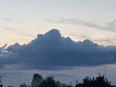 The image depicts a cloudy sky over a rural landscape. In the foreground, silhouetted trees and utility poles with wires are visible against the muted light. The sky is dominated by large, dark clouds, with lighter, wispy clouds above. The overall scene suggests either dawn or dusk. The location is identified as Ярэміцкі сельскі Савет, Belarus. There are no people or discernible actions in the image.