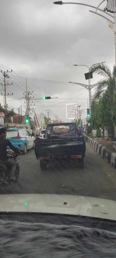 The image captures a street scene in Kutalhoksukon, Indonesia, from the perspective of a vehicle stopped in traffic. The sky is overcast and grey, suggesting a cloudy day.

In the mid-ground, a black pickup truck with a visible license plate "BA 5513 DT" is central, facing away from the viewer. Behind it, a white sedan is partially visible. To the left of the pickup, a light blue and white Mitsubishi Xpander MPV is also seen in traffic. In the immediate foreground on the left, a person on a motorcycle is partially visible, riding ahead of the Xpander. The very bottom of the image shows the dashboard or hood of the car from which the photo was taken.

The road appears to be made of asphalt or a similar material, with distinct black and white striped curbs along the right side, separating the road from a sidewalk or median with trees. Three green traffic lights are visible, indicating that traffic may be proceeding or waiting at a busy intersection.

Numerous utility poles with a dense network of overhead wires crisscross the scene, especially on the left side. Several streetlights are present; some are ornate with decorative elements, and one appears to have a small solar panel attached. Trees and other green foliage line the right side of the road and are scattered in the background. An empty billboard frame is visible in the middle distance. A sign with "10'" is partially visible on the left, near the utility poles. The overall setting is an urban or semi-urban environment with typical street infrastructure.