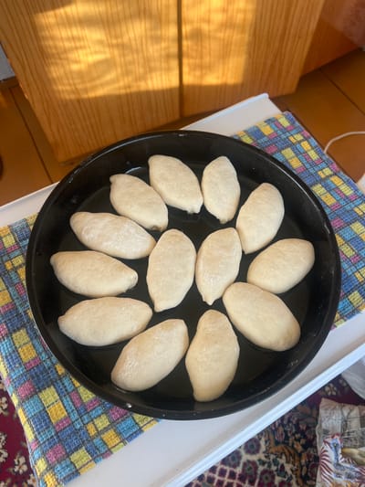The image shows a black baking pan filled with uncooked dough, arranged in a circular pattern. The dough pieces are oval-shaped and appear to be dusted with flour. The pan is resting on a colorful checkered cloth, which in turn is on a white surface. In the background, there's a wooden surface with visible grain and sunlight casting shadows, suggesting a daytime setting. A portion of a patterned rug is also visible. There is no visible text in the image. The scene appears to be a domestic setting, likely in a kitchen, where someone is preparing food. The lighting suggests it is daytime, possibly morning or afternoon, with sunlight streaming in.