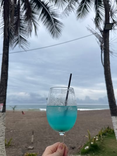 In Jacó, Costa Rica, a close-up shot focuses on a hand holding a stemmed glass filled with a bright blue cocktail. Ice cubes and a black straw are visible within the drink. The glass is covered in condensation, indicating it is cold. In the background, a sandy beach stretches towards the ocean, with gentle waves lapping the shore under an overcast sky. Two palm trees frame the scene, their fronds blurring slightly due to motion. A light fixture is attached to the trunk of the right palm tree. In the distance on the beach, a person is seated.