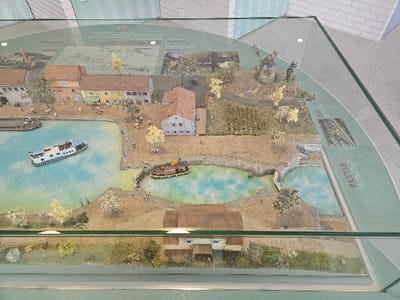 This image depicts a detailed miniature scale model displayed under a clear glass panel, representing a segment of the city of Avignonet-Lauragais, France. The model primarily showcases a canal system with associated structures and landscape features.
The central element is a light blue canal, bifurcating into a wider section and a narrower channel that connects to two smaller, interconnected water basins, resembling a lock system. A long, light blue and white leisure boat or barge occupies the wider canal section. A smaller, yellowish-brown boat is positioned within the narrower channel leading to the basins. Two narrow bridges span the connections between these smaller water bodies.
Along the main canal, a row of multi-story buildings with varying facades (tan, yellow, grey) and red-tiled roofs are depicted, some with awnings. Another large, brown-roofed building is situated in the foreground, and additional structures are scattered throughout. In the upper right quadrant, a traditional windmill stands on an elevated, grassy mound, adjacent to cultivated fields and dirt paths. Miniature deciduous trees with yellowish foliage are present across the landscape, which also features earthen banks and grassy areas.
Numerous small, multi-colored human figures are distributed throughout the model, appearing to walk along paths, congregate near buildings, and cross the bridges. The display includes informational graphics, maps, and text labels beneath the glass, with "Avignonet-Lauragais, France" and "BAJOLYER" visible along the green perimeter.