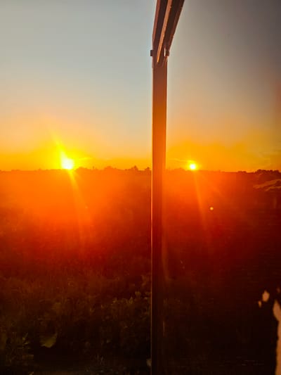 The image depicts a rural landscape in the city of Kurvovo, Russia, during sunset. Two suns are visible in the sky, creating a surreal and unusual scene. The sky transitions from a light blue at the top to a vibrant orange and yellow near the horizon. The landscape below is a field with trees silhouetted against the bright sky. A dark, vertical post, possibly part of a structure, bisects the image from top to bottom, casting a shadow and obscuring a portion of the view. The foreground is filled with dark vegetation, with some areas appearing lighter due to the sunset's glow. Lens flares are present, enhancing the intensity of the light.