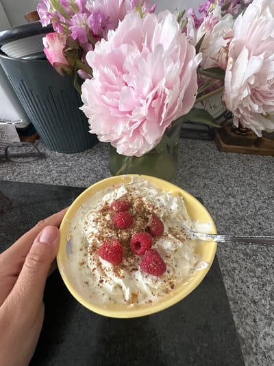 This media file captures a close-up, overhead shot of a yellow bowl filled with a creamy dessert, likely yogurt or ice cream, topped with fresh raspberries and a sprinkle of crushed nuts or granola. A spoon is partially submerged in the dessert, indicating it is being eaten or prepared to be eaten. To the left of the bowl, a bouquet of large, pale pink peonies is arranged, adding a touch of natural beauty to the scene. The background appears to be a speckled gray countertop, characteristic of a kitchen or dining area. A hand with painted nails is visible in the lower right corner, holding the bowl.
The overall impression is one of a pleasant, possibly indulgent moment, perhaps a breakfast or a light dessert. The natural light suggests it could be daytime, and the focus on food and flowers creates a calm and aesthetically pleasing atmosphere. No specific time of day or weather conditions are apparent. The location context of Macclesfield, United Kingdom, is provided externally and cannot be deduced from the image itself. There is no visible text in the media file.