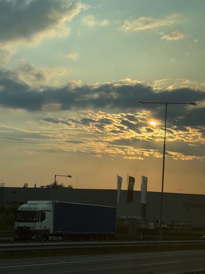 In the city of Kuří, Czechia, a truck with "SPUTNIK 96" branding is traveling on a highway during sunset. The sky is filled with dramatic clouds illuminated by the setting sun, creating a warm, golden glow. A tall street lamp stands on the side of the road. In the background, a large, dark building is visible with several flags in front of it. One flag appears to have the word "AGRO" on it, suggesting a business or agricultural-related establishment. The scene captures a moment of travel and commerce against a beautiful natural backdrop.