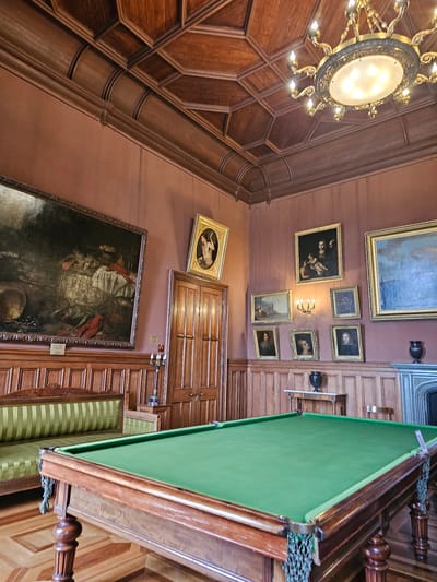 The image captures an interior space with a mix of architectural features, furnishings, and decorations, presumably in Алупкинский городской совет, Ukraine. A striking chandelier hangs from a wooden paneled ceiling. A collection of framed paintings decorates the walls. A billiard table with a green cloth top dominates the foreground. Other objects include a wooden paneled door and a chair. The photograph is taken at an angle, showcasing the height and depth of the room.