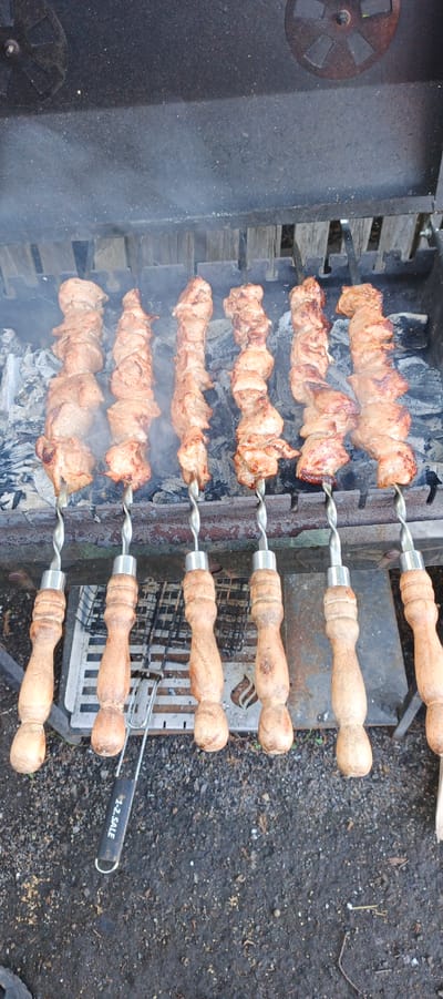 The image shows eight skewers of meat being grilled over hot coals in a brazier. The meat appears to be pork, cut into bite-sized pieces and skewered tightly. Smoke rises from the coals and the meat, indicating it is actively cooking. The skewers have decorative, twisted metal shafts and wooden handles. The brazier itself is made of dark metal with an adjustable vent on the top left. Below the brazier, a metal grill rack is visible. The ground beneath the brazier is covered in dark soil, gravel, and some dry leaves, suggesting an outdoor setting. There is a tool with the text "1-2. SALE" written on its handle lying on the ground in front of the skewers. The lighting suggests it is daytime, and there are no visual cues about the weather, though the smoke indicates it is likely not raining. The scene depicts a common outdoor cooking activity, likely a barbecue or shashlik preparation, given the context of Petukhovo, Russia, where such dishes are popular.
