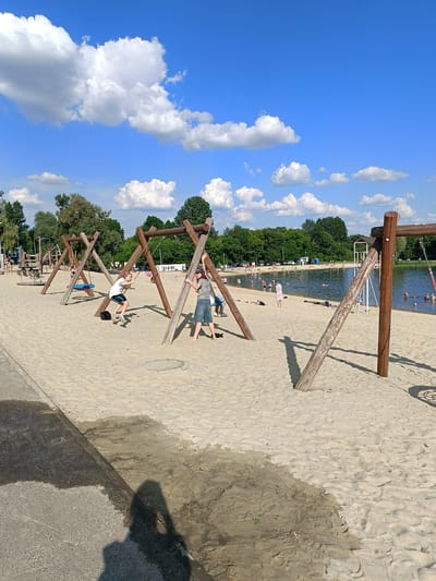The image depicts a sunny day at a sandy beach area adjacent to a body of water, likely a lake or reservoir, with a backdrop of green trees and a bright blue sky dotted with white clouds. In the foreground, several wooden swing sets are visible on the sand. Two children are actively using the swings, one girl in a white shirt and dark shorts is mid-swing, while another child, a boy in a grey shirt and denim shorts, stands near a swing set, appearing to hold onto one of its wooden supports.

Further into the scene, the water is populated with numerous people swimming, wading, and relaxing. Some are floating on the water, while others are closer to the shore. Beach chairs and towels are scattered on the sandy area near the water's edge. In the distance, there are more trees and a parking lot with cars, suggesting this is a recreational area. The sand shows footprints and some damp patches, indicating recent activity and moisture. The overall impression is of a lively public beach on a warm, summer day.

This location is the beach at the Gubkin Reservoir in Gubkin, Russia.