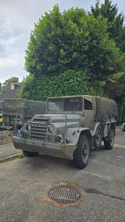 An olive-drab, six-wheeled military truck, possibly a DAF, is parked on an asphalt surface in front of a row of houses and dense green trees. The truck has a canvas covering over its cargo area, which is draped with camouflage netting. The front of the truck features a large grille, a license plate reading "BE 12-14," and a DAF logo. A manhole cover with a grate pattern is visible on the road in the foreground. The sky is overcast with some clouds. The scene appears to be in Zeddam, Netherlands, based on a partial inscription on the manhole cover that reads "VANRINGBOO."