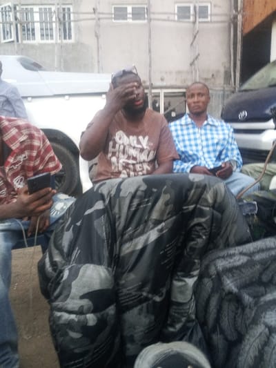 Three men are seated outdoors. One wears a brown shirt with "Only Human After All" printed on it and sunglasses perched on his head, covering his eye with his hand. Another man wears a blue and white plaid shirt. A third person in a red plaid shirt holds a phone. Large, dark camouflage bags are in the foreground, obscuring some of the scene. Vehicles are visible in the background, along with a building with white-framed windows.