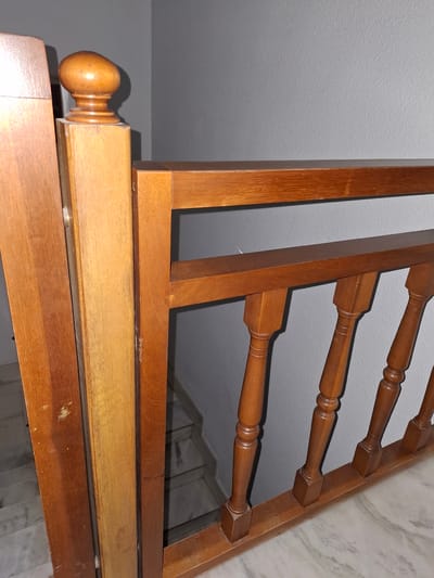 The image shows a portion of a wooden bed frame in an indoor setting. The frame consists of several horizontal rails connected to vertical posts, forming a headboard or footboard. The wood is finished with a glossy varnish and has a warm, brown tone. The backdrop includes a plain white ceiling and a section of marble or patterned wall, indicating the bed is located against a wall. I have no knowledge of the city Xirivella or where the image would be located there.