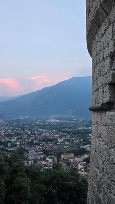 The image shows a scenic view from a high vantage point, likely a castle or fortress, overlooking a town nestled in a valley. The foreground features a textured stone wall, possibly part of the fortification, with a protruding cylindrical object, perhaps a remnant of a structure or decoration.

Below the stone structure, the scene opens up to a breathtaking panorama. A vast, hazy blue mountain range dominates the left side of the frame, its slopes softened by distance and the atmospheric conditions. A portion of the sky on the far left displays a gentle gradient of pale blue transitioning into soft pink hues, suggesting either sunrise or sunset.

To the right of the mountain, a sprawling town is visible, characterized by a dense collection of buildings with terracotta roofs and pastel-colored facades. The urban landscape stretches out into the valley, interspersed with patches of lush green vegetation. The town appears to be illuminated, with some windows showing lights, indicating either early morning or evening.

The right edge of the image is framed by the dense, dark green foliage of trees, providing a natural border to the view. The overall atmosphere is serene and picturesque, conveying a sense of historical grandeur combined with natural beauty. The time of day is likely dusk or dawn, given the sky colors and the lighting in the town. The location cues, such as the architecture and the surrounding mountainous terrain, are consistent with Riva del Garda, Italy. No text is visible in the image.