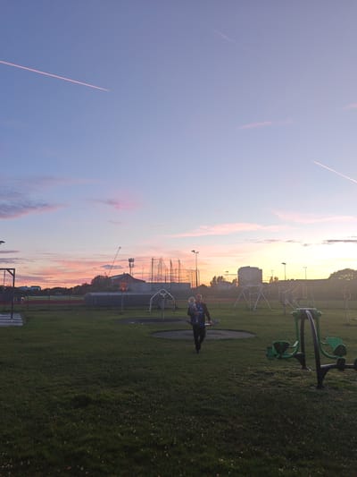 A man is walking across a grassy field, holding a baby. He is wearing a dark blue shirt with a white logo and black pants. The baby is wearing a light-colored outfit. In the background, there is a playground with swings and other equipment, as well as some buildings and a crane. The sky is a gradient of blue, pink, and orange, with two contrails from airplanes visible. The image appears to have been taken at sunset. The location is likely a public park or recreational area in Worthing, United Kingdom, given the presence of playground equipment and the distinct contrail patterns often seen in the sky over populated areas. The overall scene suggests a peaceful evening outing.
