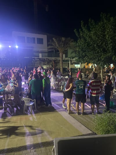 The image depicts an outdoor nighttime gathering in Blanca, Spain. A crowd of people are standing around tables and chairs, some holding drinks, suggesting a social event or party. A stage with bright lights is visible on the left side of the frame, indicating a possible performance or DJ set. The background shows buildings with windows and balconies, and trees, including a palm tree, are present in the scene, suggesting a warm climate. A crane is also visible in the distance, hinting at ongoing construction or development. The overall atmosphere appears lively and casual.