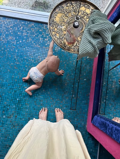 A baby in a diaper is crawling on a blue-tiled floor. A person's feet and white dress are visible near the baby. An ornate gold-colored table with a green towel and diffuser sits to the left of the baby. A mirror with a blue and pink frame is in the upper portion of the image. The scene is indoors. There is no visual information to support this scene being in Чёрное, Russia.