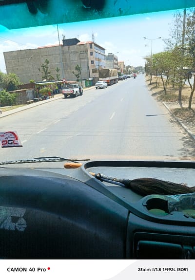 The image captures a forward-facing view from inside a vehicle onto a street in Viwandani ward, Kenya, under bright, clear daylight. The lower third of the frame shows a dark dashboard, featuring a light brown, cylindrical object and a dark, tasseled item on its surface. A white sticker with red and black text, including "DGC," is affixed to the windshield on the lower left.
The asphalt road, marked with white lines, extends straight into the distance, appearing to have at least two lanes. On the left side of the road, a dirt shoulder precedes a concrete sidewalk bordered by a series of small, corrugated metal structures. Several individuals are visible near these structures. A white Nissan pickup truck with a roll bar and a "PROMAN" decal on its tailgate is parked on this shoulder. Beyond these foreground elements, a large, multi-story concrete building with unpainted walls dominates the midground, flanked by other multi-story residential or commercial buildings featuring varied facades and rooftop antennae.
On the right side of the road, a similar dirt shoulder and concrete sidewalk are lined with evenly spaced young trees displaying sparse green foliage. Streetlights are visible along this side. Vehicles, including a long truck covered with a tan tarp, a white sedan, and numerous other cars and motorcycles, are observed traveling in the same direction, receding into the background. Distinct shadows are cast on the road and sidewalks, indicating a high sun angle. The sky above is clear blue with minimal cloud cover.