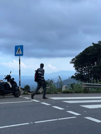 A medium-shot photo captures a scene near Seetha Eliya, Sri Lanka. An individual in casual clothing crosses a marked zebra crossing on a paved road. The person has a backpack. A black motorcycle is parked on the road's edge, close to a blue pedestrian crossing signpost. A backdrop of layered mountains fades into a hazy sky. Vegetation lines the roadside, and the sky is overcast.