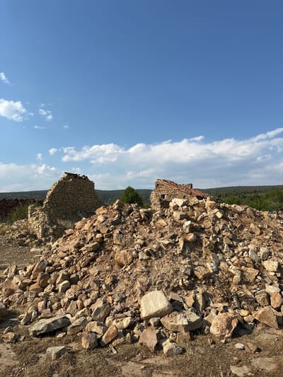 A low-angle, outdoor, full shot captures the ruins of two stone structures against a bright blue sky dotted with white clouds. In the foreground, a large, disordered pile of rocks, varying in size and color from light tan to reddish-brown, dominates the lower half of the frame. Some dry grass and small weeds are visible amongst the stones. Behind this rubble pile, the partially collapsed remains of two stone buildings are visible. The structure on the left is a tall, thick wall of rough-hewn stones, with a section of weathered red roof tiles precariously balanced on top. To the right, another building fragment shows a similar stone construction with a steeply angled, partly intact red tile roof. Small windows or openings are visible in this structure. In the middle ground, a dense line of green trees and vegetation stretches across the horizon, suggesting a wooded area or hillside. A single, larger green tree stands prominently behind the rightmost ruin. In the far background, a distant, undulating line of hills fades into the background. The overall impression is one of decay and abandonment in a rural, possibly Mediterranean, landscape. The scene is likely located in Alpuente, Spain, given the architectural style and the characteristic landscape.