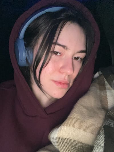 A person lies reclined, wearing a maroon hooded sweatshirt. Headphones are visible beneath the hood. Hair frames the face, partially obscuring the left eye. The person gazes directly at the camera with a neutral expression. A tan and brown plaid blanket rests partially over the right side of the frame, adding texture and warmth to the scene. The overall ambiance suggests comfort and relaxation.