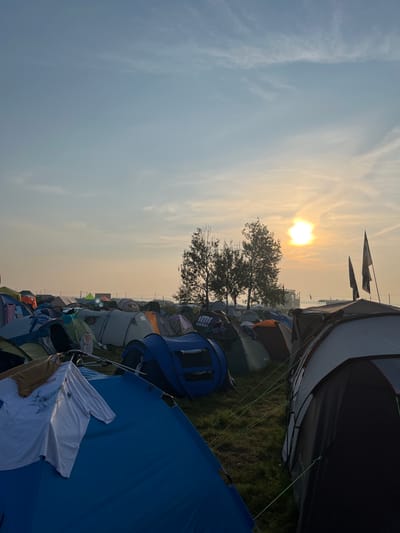 The image depicts a large campsite with numerous tents set up on a grassy field. The sun is rising or setting, casting a warm glow and creating a hazy atmosphere with some clouds in the sky. There are several trees in the mid-ground, and in the distance, there are some structures and possibly a body of water. The foreground shows a close-up of several tents, with some clothes hanging on a tent pole in the lower left. The scene appears to be a temporary setup, likely for an event or festival, given the density and variety of the tents. The location is identified as Surakovo, Russia.