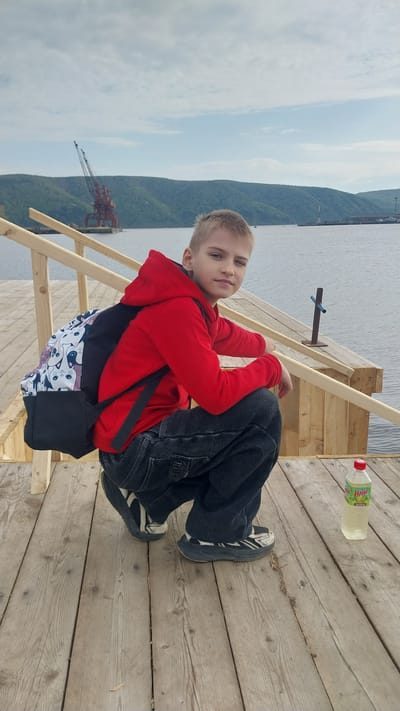 The image features a young boy, approximately 10-14 years old, with short blonde hair and fair skin, squatting on a wooden pier in Nikolayevsk-on-Amur, Russia. He is wearing a red hooded sweatshirt, dark wide-leg jeans, and dark athletic shoes with white accents. A black backpack with a white upper section patterned with cartoon cat faces (white, grey, black) is visible on his back. The boy is looking directly at the viewer, with his right eye slightly narrowed. To his right, on the wooden pier, a plastic bottle with a red cap and a green label (partially visible as "Напиток") contains a light-colored liquid. The pier consists of light brown, weathered wooden planks and features a wooden railing structure behind the boy. In the midground, a calm body of water, likely the Amur River or a bay, stretches across the scene under a partially cloudy sky. The background reveals green, tree-covered hills or mountains on the opposite shore. To the far left, a large, rusty red industrial crane is visible on the distant land. Further to the right in the background, additional industrial structures are present on the distant riverside.