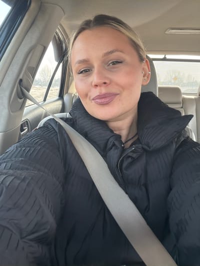 An adult female, appearing to have light blonde hair pulled back, is depicted in a close-up selfie taken from the passenger seat of a vehicle. She wears a black puffer jacket with a textured, pleated design and a light-colored seatbelt extending diagonally from her left shoulder across her chest. A dark beaded necklace is visible around her neck. Her left ear displays multiple piercings. Her facial expression is a slight smile, with visible makeup including defined eyebrows, mascara, and medium-toned lipstick. The vehicle's interior features light-colored upholstery and door panels. Through the car's passenger window, an exterior scene of an undeveloped area is visible, characterized by bare trees and exposed ground under natural daylight. This scene is located in the city of Дулесово, Russia.