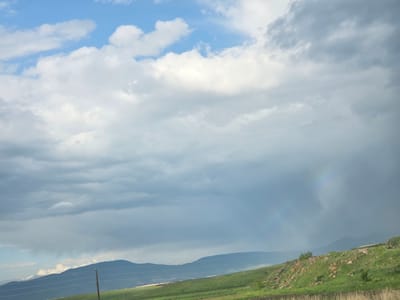 The image shows a landscape with rolling hills under a cloudy sky. The sky is a mix of blue and white clouds, with some areas appearing dark and heavy, suggesting an impending or ongoing rain shower. A faint rainbow is visible on the right side of the frame. The foreground consists of green grass-covered hills with scattered rocks. In the mid-ground, there are hills with a more defined slope, and a single wooden pole with wires stretches across the scene. The background features a mountain range silhouetted against the sky. The overall impression is of a rural or mountainous area. This scene is likely located in the vicinity of Azatan, Armenia, given the geographical context. There are no people or animals visible in the image.
