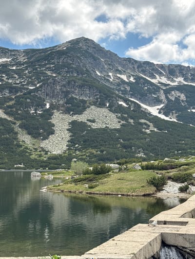 This is a vertical, eye-level shot of a mountain lake with a rugged mountain range in the background. The lake occupies the lower left portion of the image and reflects the cloudy sky and surrounding mountains. The shore of the lake in the mid-ground is covered in grass and low-lying green bushes, with scattered rocks. The mountain slopes are a mix of dark green coniferous trees, rocky scree, and patches of snow. The sky is partly cloudy with dramatic white clouds against a blue sky. In the foreground, there is a concrete structure with water flowing over its edge on the right side. This scene is located in the Rila Mountains near Avramovo, Bulgaria, a region known for its glacial lakes and alpine scenery. There are no people or animals visible in the image.