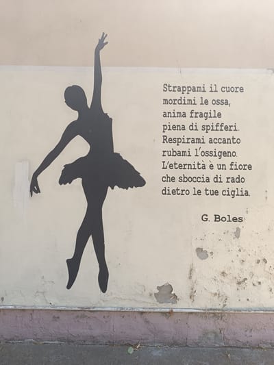 The image shows a black silhouette of a ballet dancer in mid-pose on a weathered, light-colored wall. The dancer's left arm is extended upwards, and her right arm is bent at the elbow with her hand reaching downwards. She is wearing a tutu. To the right of the dancer is a poem written in a typewriter font in black ink. The poem reads:

Strappami il cuore
mordimi le ossa,
anima fragile
piena di spifferi.
Respirami accanto
rubami l'ossigeno.
L'eternità è un fiore
che sboccia di rado
dietro le tue ciglia.

Below the poem is a signature: "G. Boles". The wall itself shows signs of age and wear, with peeling paint and cracks. A white pipe runs horizontally across the lower section of the wall, above a band of pink-painted masonry. The ground in front of the wall is a dark, rough surface. The location is identified as Casalmaggiore, Italy, specifically on a wall near the "Ponte sul Po" (Po River Bridge).
