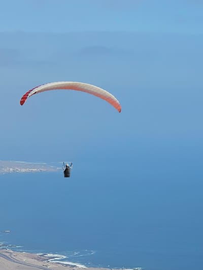 The image shows a paraglider soaring through a clear blue sky in Alto Hospicio, Chile. The paraglider has a red, white, and blue canopy. In the distance, the coastline and terrain of Alto Hospicio are visible, including the road along the beach. A person is seen in a wing suit flying near the paraglider.
