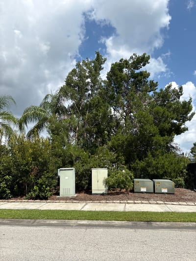 In Parrish, Florida, along a paved road bordered by a strip of green grass, a sidewalk runs parallel to a landscaped area. This landscaped area is populated by lush green bushes and trees, with a prominent cluster of tall, mature trees forming a backdrop. In front of the trees, and on the landscaped strip, stand three utility boxes. Two of the boxes are identical in size and olive-green in color, each displaying yellow warning labels and numbers in the upper center. The third box, to the left of these two, is taller and a lighter green, also featuring a warning label. The sky above is a mix of bright blue with scattered white and gray clouds.