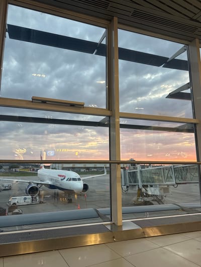 A British Airways airplane is parked at the gate at Otopeni Airport in Otopeni, Romania, as seen from inside the terminal. The plane is white with the British Airways logo on its side. A jet bridge is connected to the aircraft. In the background, the sky is filled with dramatic clouds at sunset, casting a warm orange glow. The airport terminal's large windows offer a view of the tarmac and the airplane. The scene appears to be during the golden hour, with the setting sun creating a vibrant atmosphere.