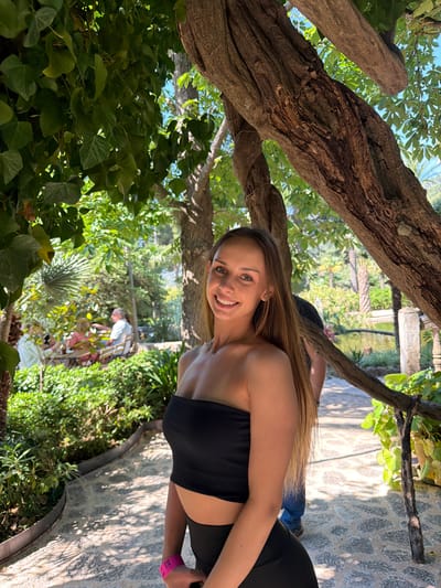 A medium-shot photo shows a woman in a black bandeau top and black leggings smiling and looking towards the camera. She appears to be standing near a tree with thick, gnarled branches, surrounded by lush vegetation. In the background, there is a glimpse of a garden scene with stone pathways and other people. The location may be in Bunyola, Spain, given the presence of stone, a garden, and local vegetation.