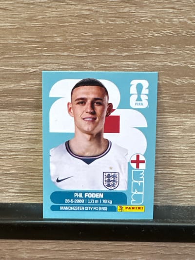 An England football sticker featuring Phil Foden is displayed on a wooden surface. The sticker has a light blue background with a large white graphic of the number '2' and part of a '3'. Phil Foden is depicted in the center wearing an England jersey. Below his image, text reads "PHIL FODEN", "28-5-2000 | 1,71 m | 70 kg", and "MANCHESTER CITY FC (ENG)". The Panini logo is visible in the bottom right corner. The scene is static with no actions or interactions occurring. The environment is a close-up view of the sticker on a textured wooden surface. No elements in the image suggest the city of Prisovo, Bulgaria.